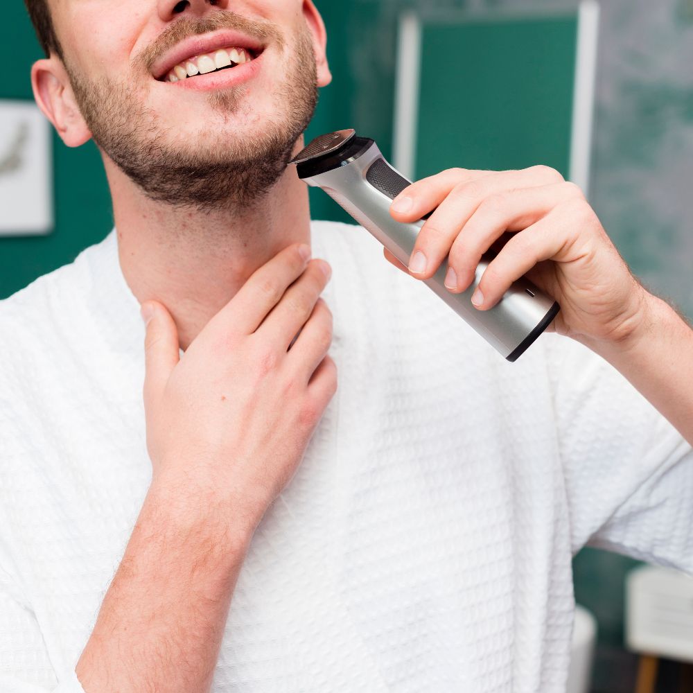 Man using an electric razor on his beard with a towel around his neck.