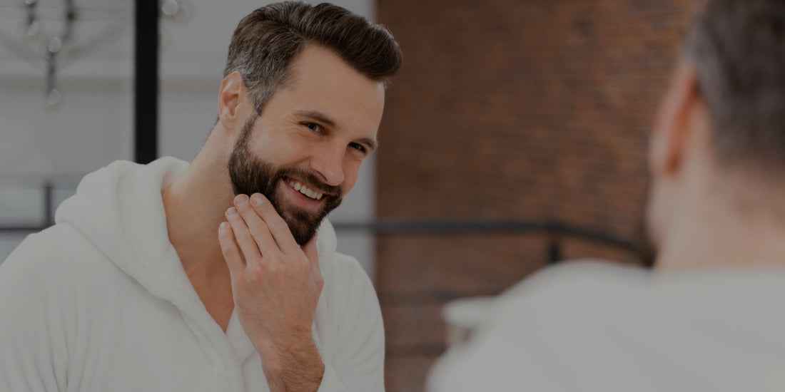 Man with a beard smiling and touching his face in front of a mirror.