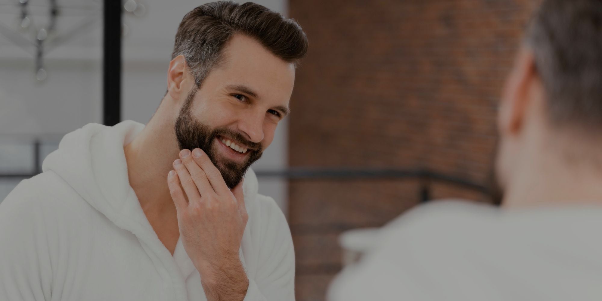 Man with a beard smiling and touching his face in front of a mirror.