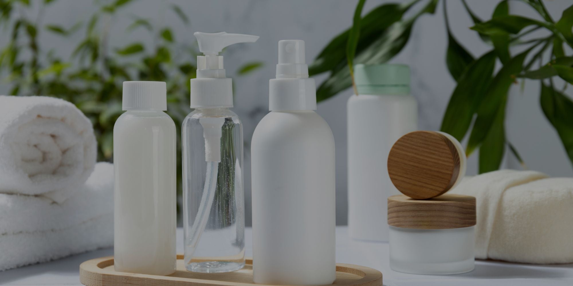 Set of skincare products including bottles and jars on a wooden tray with a blurred green plant background.