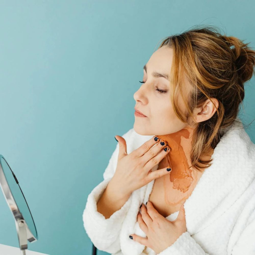 Woman applying a brown paste to her neck against a light blue background

